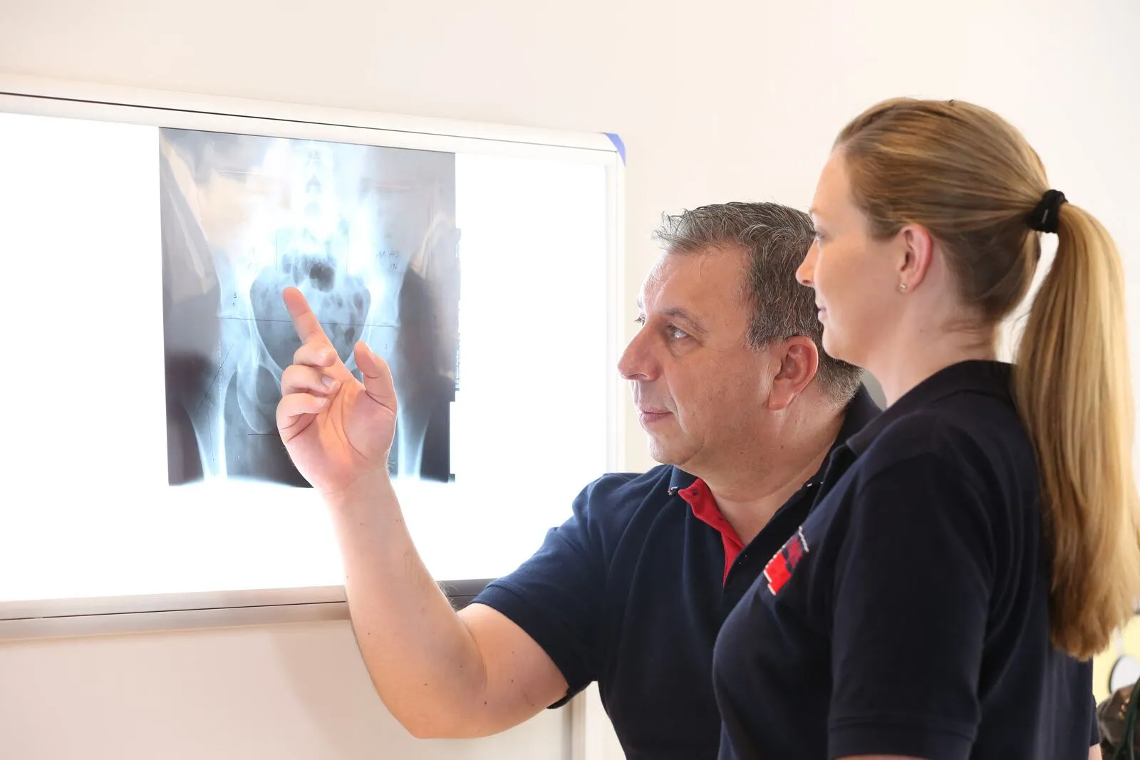 Arzt zeigt Röntgenbild der Hüfte am Leuchtkasten, Patientin schaut zu